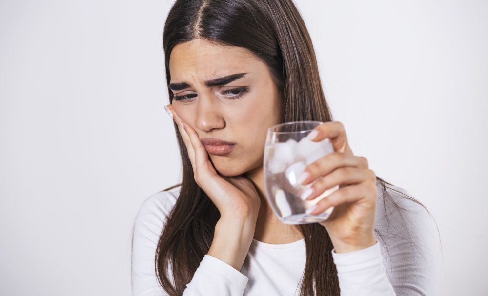 A woman drinks a glass of cold water and reacts to sudden tooth sensitivity, holding her cheek in discomfort.