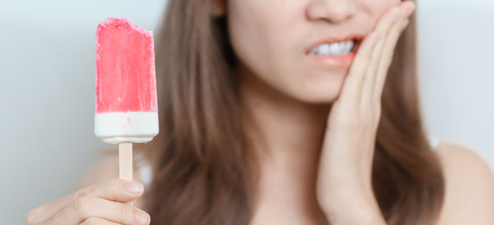 A woman touches her cheek and winces in pain, showing symptoms of tooth sensitivity or dental discomfort.